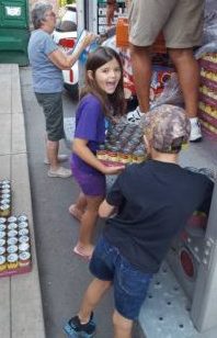 A smiling young girl loads a box filled with soup cans onto a box truck